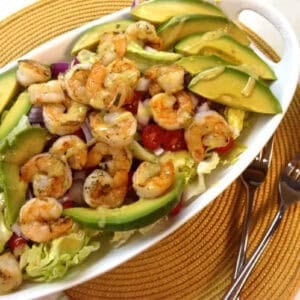 An overhead view of Grilled Shrimp and Avocado Salad with iceberg lettuce and cherry tomatoes topped with lemon mustard vinaigrette in an oval-shaped dish next to two forks and two bowls