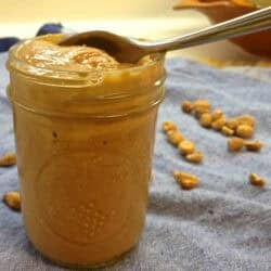 A jar of homemade peanut butter with a spoon on a blue cloth napkin with scattered peanuts