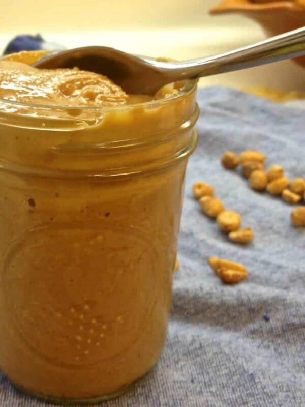 A jar of homemade peanut butter with a spoon on a blue cloth napkin with scattered peanuts