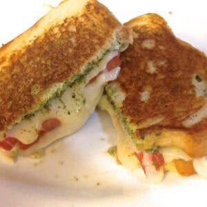 Overhead view of Grilled Fresh Mozzarella, Tomato and Pesto Sandwich cut in half with cheese oozing from the sides on a plate