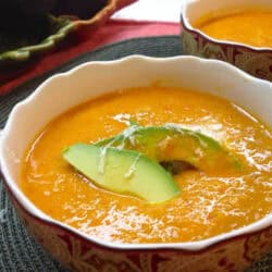 A bowl of Roasted Sweet Corn and Tomato Soup topped with two slices of avocado and grated asiago cheese next to another bowl of soup