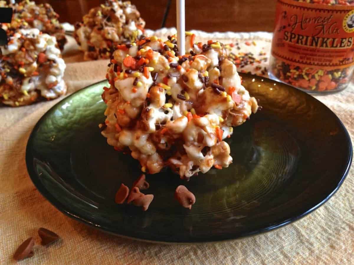 Marshmallow Popcorn Balls Zagleft A Holiday Marshmallow Popcorn Ball topped with orange, yellow, and brown fall sprinkles on a small plate with three more popcorn balls and a jar of sprinkles in the background