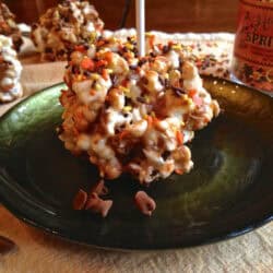 A Holiday Marshmallow Popcorn Ball topped with orange, yellow, and brown fall sprinkles on a small plate with three more popcorn balls and a jar of sprinkles in the background