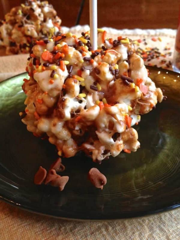 A Holiday Marshmallow Popcorn Ball topped with orange, yellow, and brown fall sprinkles on a small plate with three more popcorn balls and a jar of sprinkles in the background