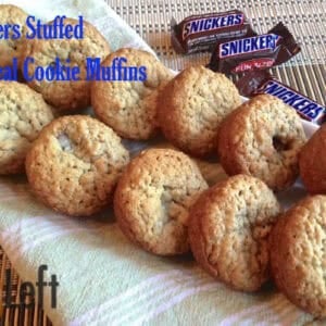 Twelve snickers stuffed oatmeal cookie muffins in a long basket lined with a green and white striped cloth napkin next to three snack-sized snickers candy bars