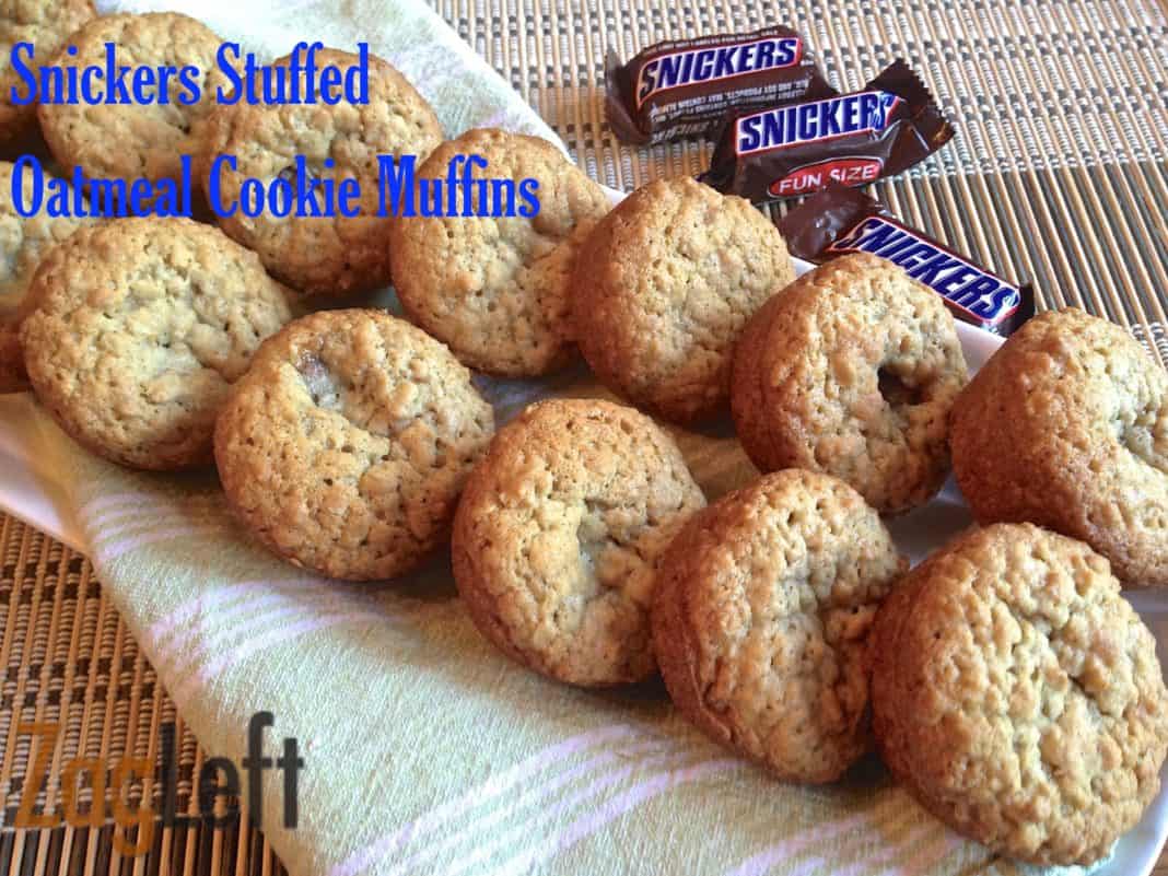 Twelve snickers stuffed oatmeal cookie muffins in a long basket lined with a green and white striped cloth napkin next to three snack-sized snickers candy bars
