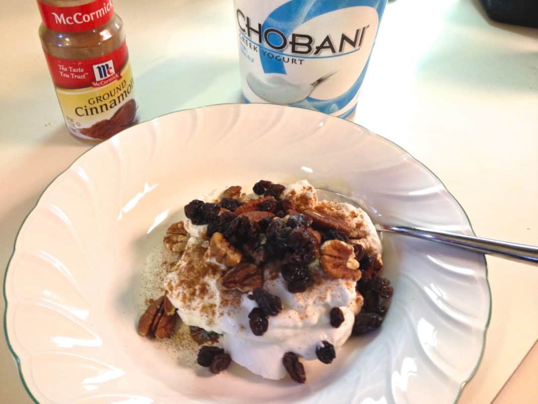 A bowl of Greek yogurt topped with cinnamon, raisins, and pecans next to a container of ground cinnamon and a container of greek yogurt 