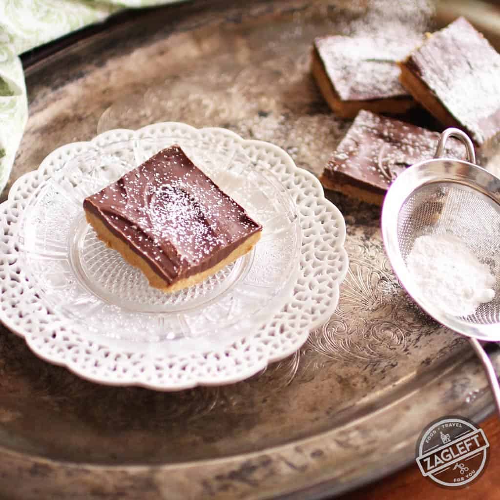 A No Bake Chocolate Peanut Butter Bar dusted with powdered sugar on a small plate next to three more bars all on a metal tray