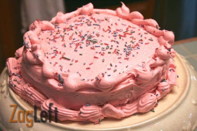 Strawberry Cake with pink icing topped with purple and pink sprinkles on a cake stand 