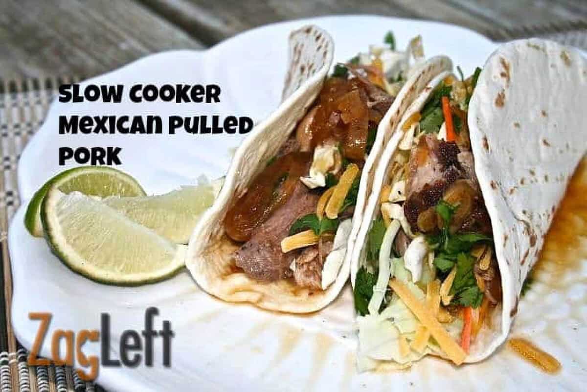 Two tacos with Slow Cooker Mexican Pulled Pork topped with shredded cabbage, chopped cilantro, a little cheese, and extra gravy in flour tortillas on a plate garnished with three lime slices
