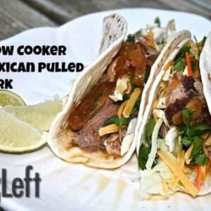 Two tacos with Slow Cooker Mexican Pulled Pork topped with shredded cabbage, chopped cilantro, a little cheese, and extra gravy in flour tortillas on a plate garnished with three lime slices