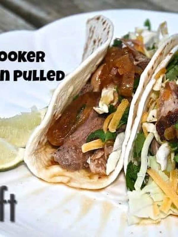 Two tacos with Slow Cooker Mexican Pulled Pork topped with shredded cabbage, chopped cilantro, a little cheese, and extra gravy in flour tortillas on a plate garnished with three lime slices
