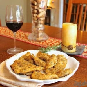 A large plate of Baked Italian Chicken Tenders garnished with a sprig of rosemary next to a glass of red wine and a candle on a wooden table