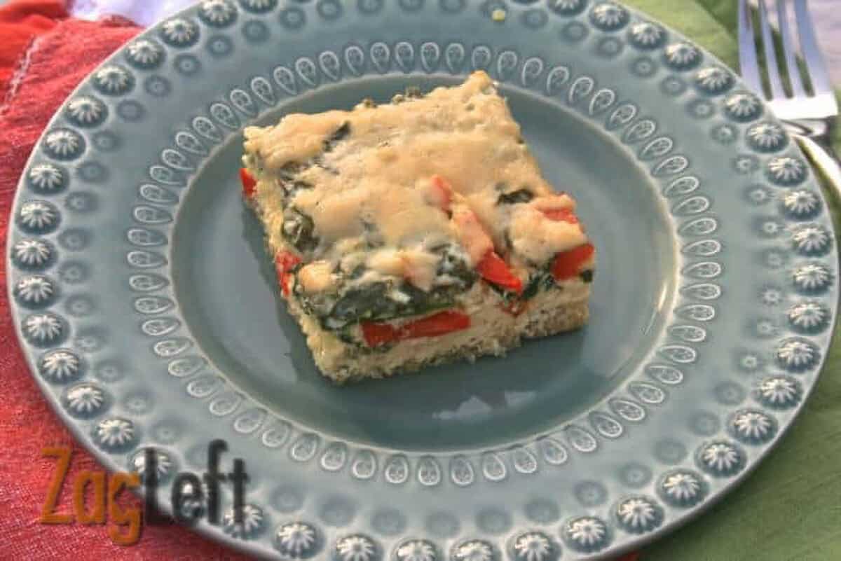 A square slice of Baked Quinoa with Spinach, Red Peppers and Cheese on a blue plate next to a fork