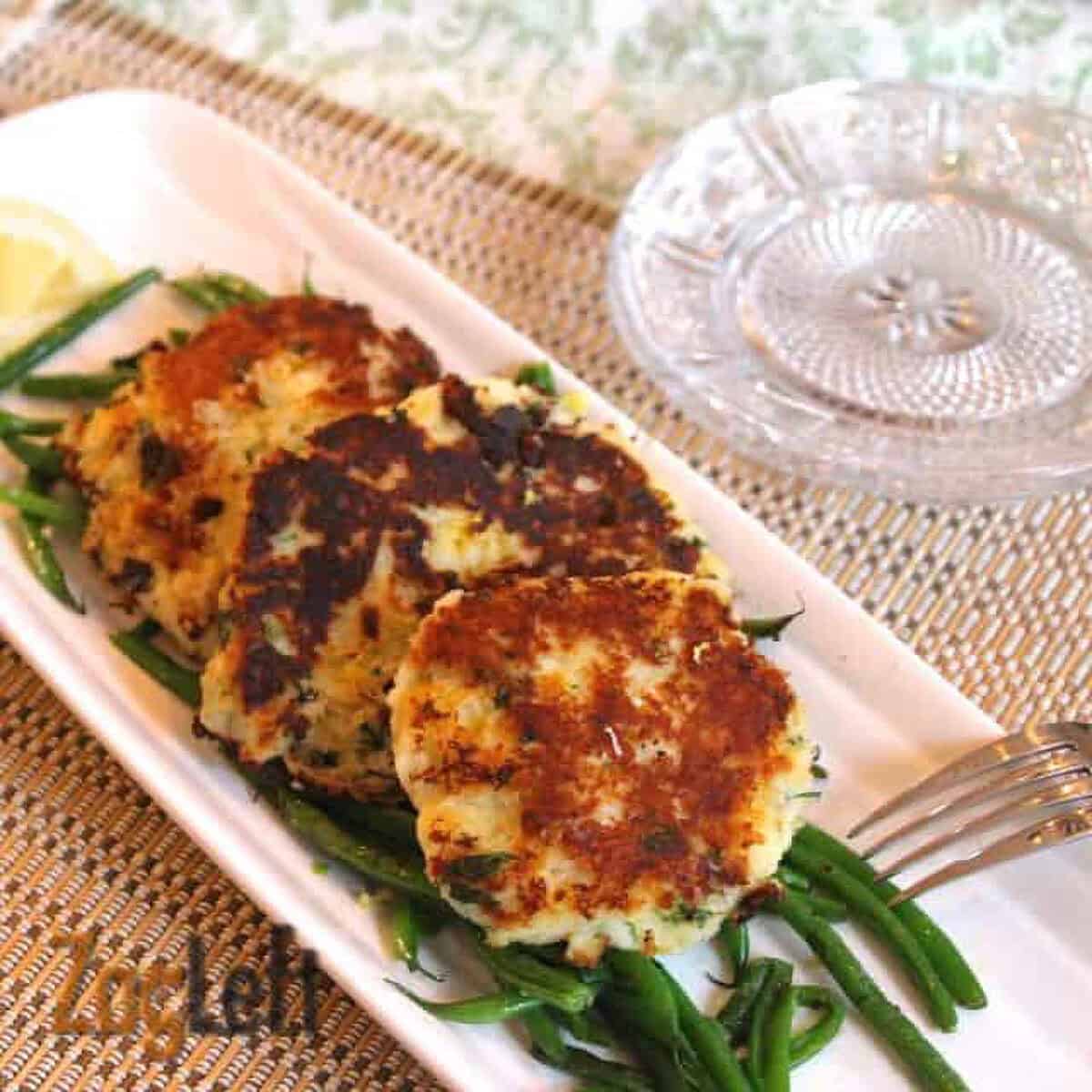 Three Parmesan Mashed Potato Patties on a bed of green beans on a long rectangle dish garnished with two lemon slices next to a stack of small plates