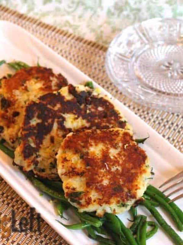 Three Parmesan Mashed Potato Patties on a bed of green beans on a long rectangle dish garnished with two lemon slices next to a stack of small plates