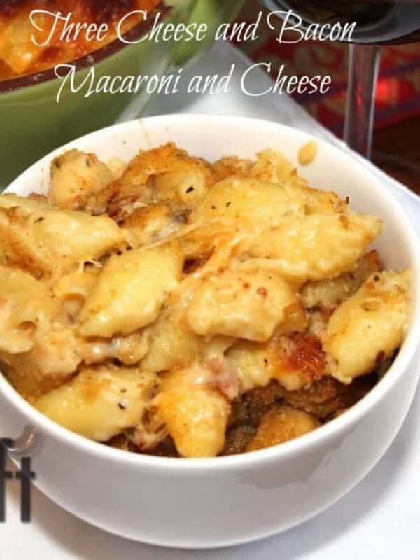 A closeup of a bowl of Three Cheese and Bacon Macaroni and Cheese plated next to a glass of red wine and a baking dish with the remainder of the macaroni and cheese