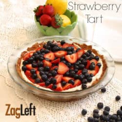 Strawberry Tart topped with strawberry slices and blueberries in a pie dish next to a handful of scattered blueberries and small bowl of two lemons and four strawberries