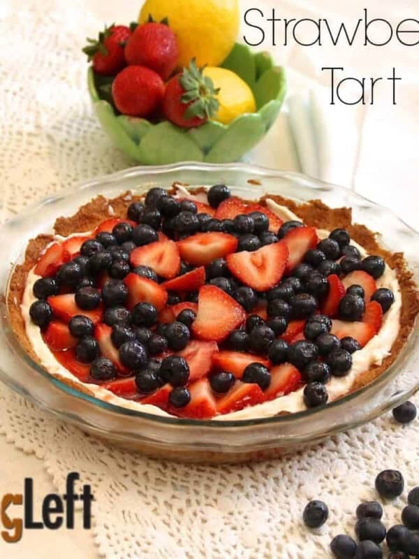 Strawberry Tart topped with strawberry slices and blueberries in a pie dish next to a handful of scattered blueberries and small bowl of two lemons and four strawberries