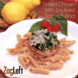 Grilled Chicken breasts on a bed of pasta topped with spinach and parmesan cheese on a plate garnished with three cherry tomatoes and two large basil leaves next to a small plate of two lemons