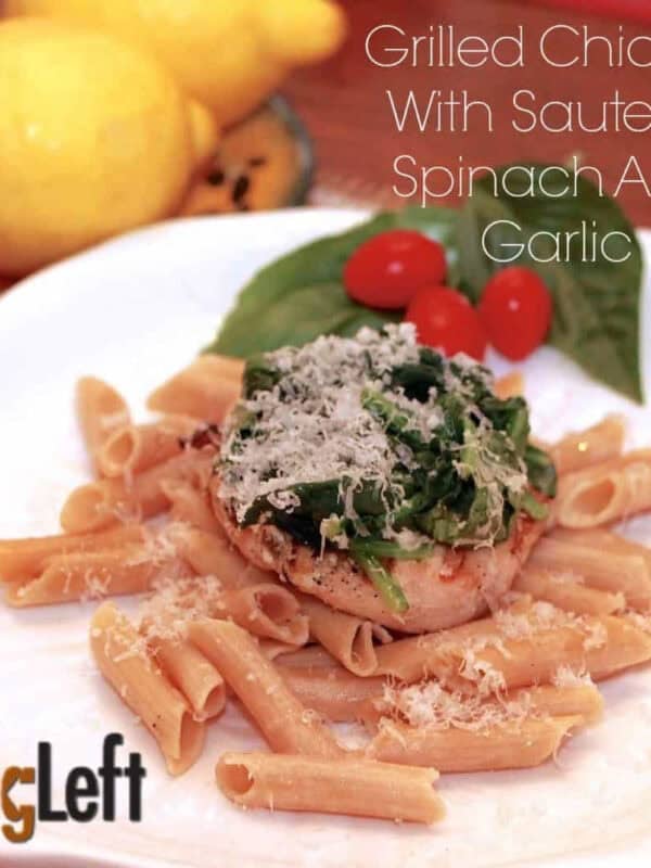 Grilled Chicken breasts on a bed of pasta topped with spinach and parmesan cheese on a plate garnished with three cherry tomatoes and two large basil leaves next to a small plate of two lemons