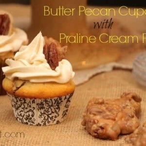 Two Butter Pecan Cupcakes with Praline Cream Filling topped with white icing, a piece of a praline, and a pecan next to two pralines on a burlap fabric