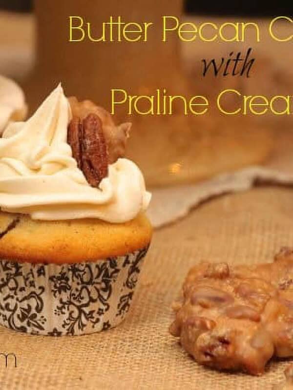 Two Butter Pecan Cupcakes with Praline Cream Filling topped with white icing, a piece of a praline, and a pecan next to two pralines on a burlap fabric