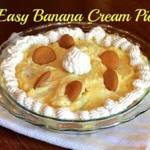 Banana Cream Pie topped with whipped cream and vanilla wafers in a glass circular pie dish on a white and yellow striped cloth napkin