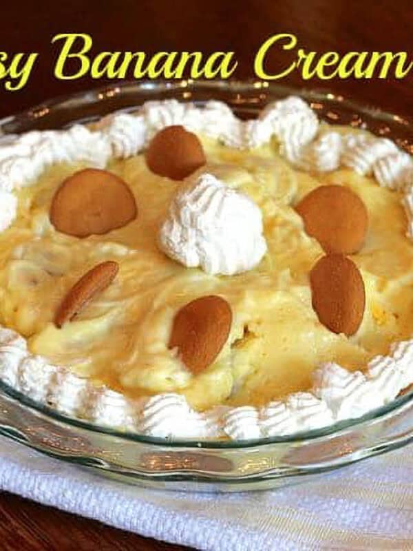 Banana Cream Pie topped with whipped cream and vanilla wafers in a glass circular pie dish on a white and yellow striped cloth napkin