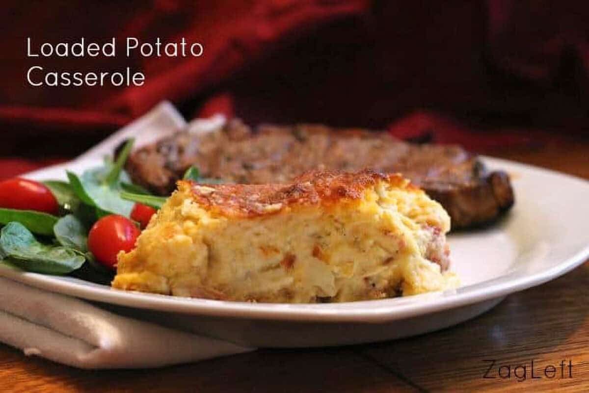 A slice of Loaded Potato Casserole on a plate with a steak and a side salad of spinach and cherry tomatoes