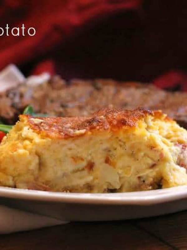 A slice of Loaded Potato Casserole on a plate with a steak and a side salad of spinach and cherry tomatoes