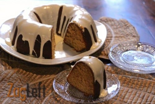 An overhead view of a slice of Eggnog Bundt Cake topped with white icing on a small plate next to the remainder of the cake on a large plate and a stack of small plates