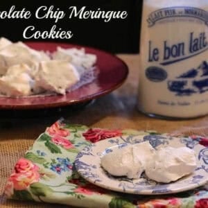 Two Chocolate Chip Meringues on a small plate on a floral cloth napkin next to a large plate of meringues and a bottle of milk