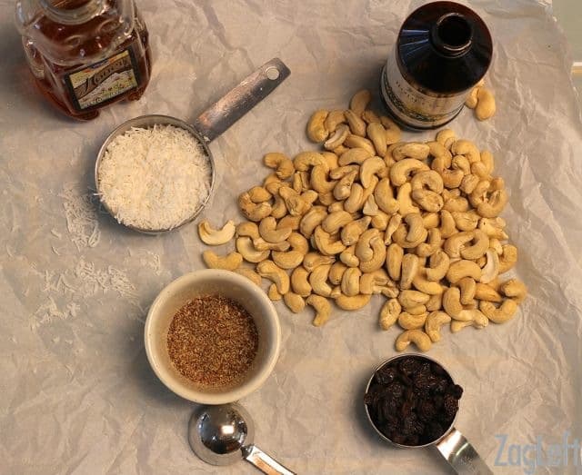 Overhead view of Ingredients- Scattered Cashews, measuring cups of unsweetened coconut, ground flaxseed, and raisins and a container of honey