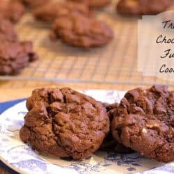 Three Triple Chocolate Fudge Cookies on a small plate on a blue cloth napkin with more cookies on a cooling rack in the background