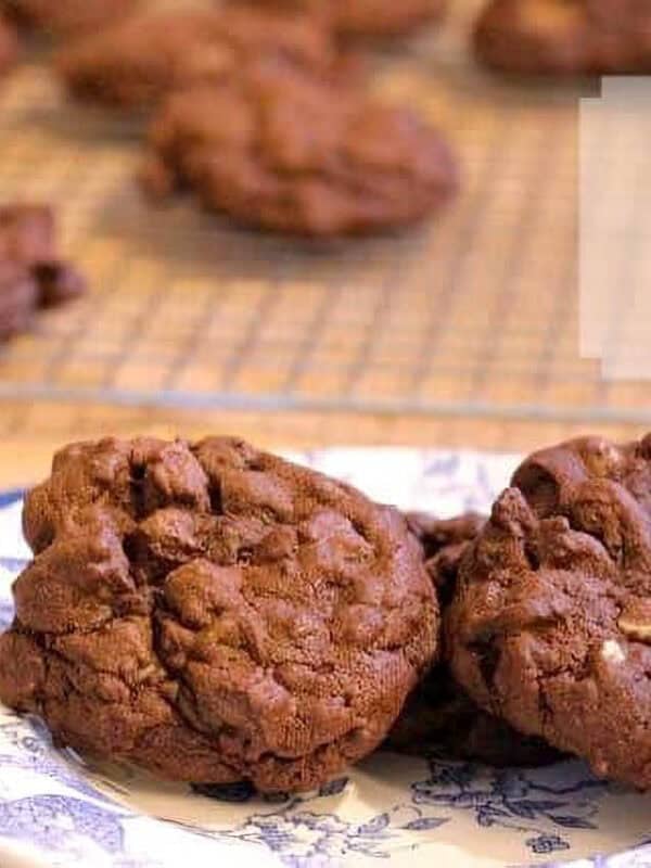 Three Triple Chocolate Fudge Cookies on a small plate on a blue cloth napkin with more cookies on a cooling rack in the background