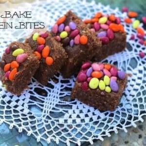 Five square No Bake Protein Bites topped with colorful chocolate covered sunflower seeds on a doily with scattered colorful chocolate covered sunflower seeds