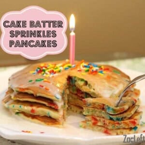 A stack of Four Birthday Cake Batter Sprinkles Pancakes topped with rainbow sprinkles, glaze, and a lit birthday candle on a plate with a forkful being taken from the plate