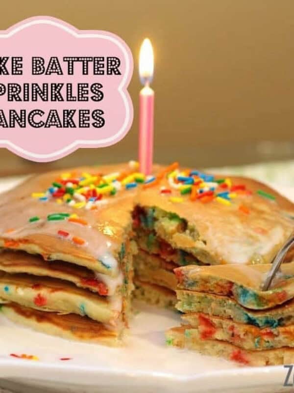 A stack of Four Birthday Cake Batter Sprinkles Pancakes topped with rainbow sprinkles, glaze, and a lit birthday candle on a plate with a forkful being taken from the plate