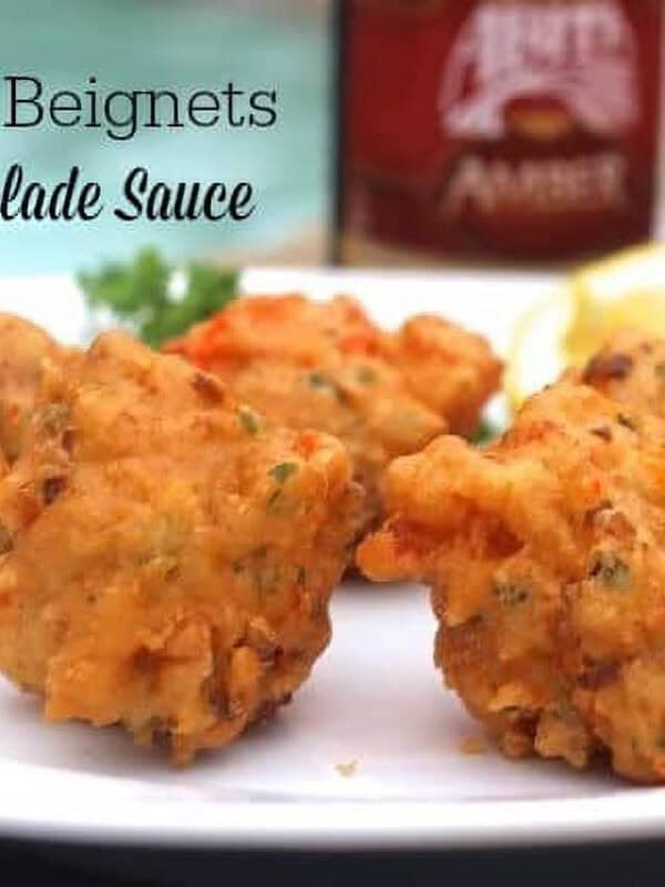 Five Crawfish Beignets on a plate with lemon slices next to a glass of Remoulade Sauce and a bottle of beer with a pool in the background