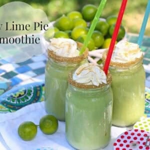 Three Key Lime Pie Smoothies with straws, crushed graham cracker crumbs on the rim, and topped with whipped cream and more graham cracker crumbs on a tray next to a bowl of key limes