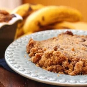 A slice of Banana Bread with a cinnamon streusel topping on a plate next to the remainder of the bread in a loaf pan and a bunch of bananas.