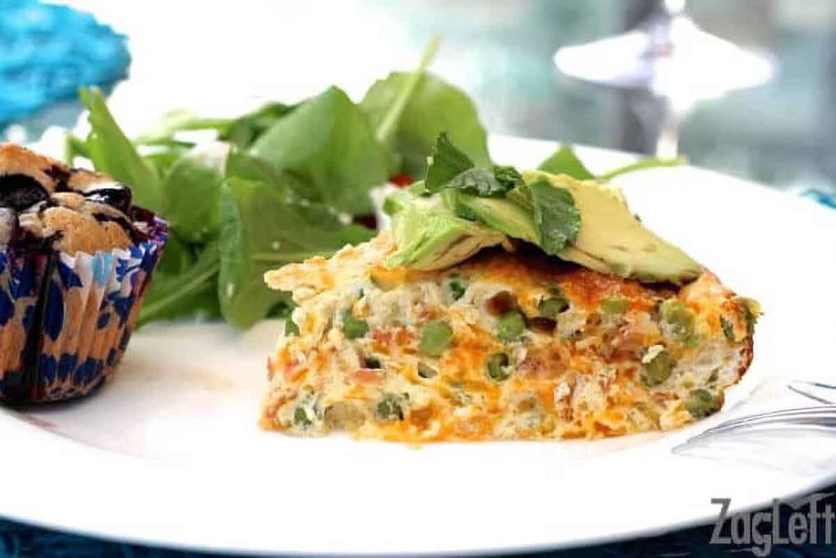 A slice of Pea, Cheddar and Crispy Pancetta Crustless Quiche topped with thin avocado slices with a spinach side salad and a blueberry muffin on a large plate