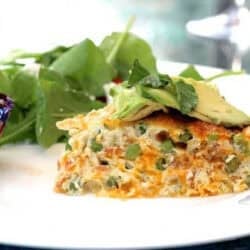 A slice of Pea, Cheddar and Crispy Pancetta Crustless Quiche topped with thin avocado slices with a spinach side salad and a blueberry muffin on a large plate