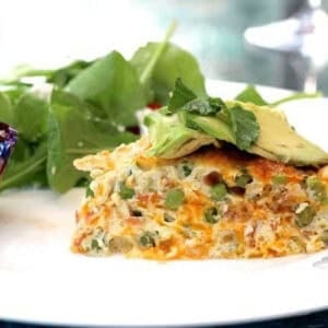 A slice of Pea, Cheddar and Crispy Pancetta Crustless Quiche topped with thin avocado slices with a spinach side salad and a blueberry muffin on a large plate