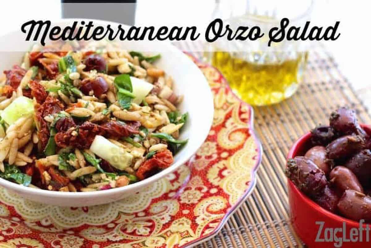 A bowl of Mediterranean Orzo Salad plated next to a glass of olive oil and a small bowl of Kalamata olives