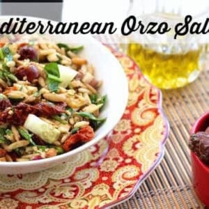 A bowl of Mediterranean Orzo Salad plated next to a glass of olive oil and a small bowl of Kalamata olives