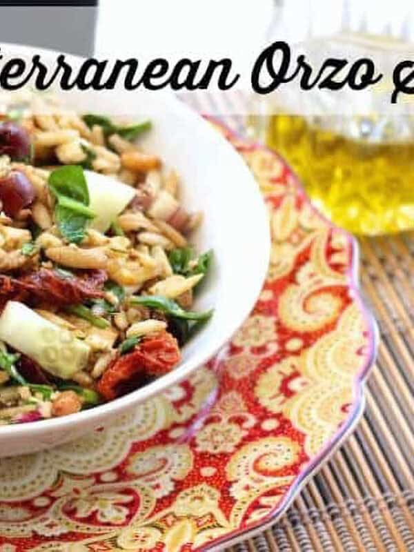 A bowl of Mediterranean Orzo Salad plated next to a glass of olive oil and a small bowl of Kalamata olives