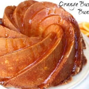 Orange Buttermilk Bundt Cake drizzled with orange glaze on a cake stand next to a small plate of orange slices.