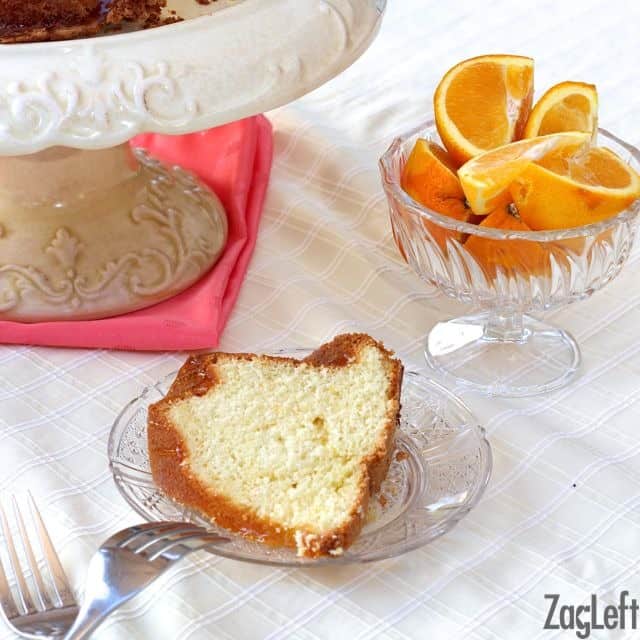A slice of Orange Buttermilk Bundt Cake on a small plate with a fork resting on the side of the plate next to a dessert glass of orange slices and a cake stand.